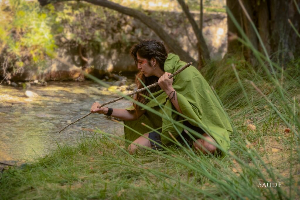Mujer con capa verde agachada junto al río con un palo, TunaTi